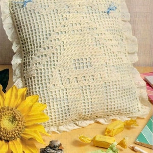 May include: A white crocheted pillow with a lacy border and blue ribbon accents. The pillow is sitting on a table with a sunflower, wrapped candies, and colored pencils.