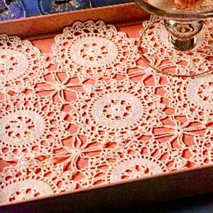 Puede incluir: Una bandeja rosa con un mantel de encaje blanco. El mantel tiene un delicado patrón floral con múltiples diseños circulares.