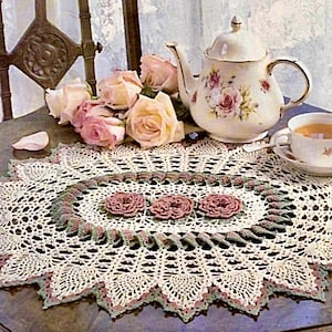 Könnte beinhalten: Eine gehäkelte ovale Tischdecke mit einem zarten Spitzenmuster und einem Mittelmotiv aus drei rosafarbenen Rosen. Die Tischdecke liegt auf einem Holztisch mit einer Teekanne und einer Teetasse.