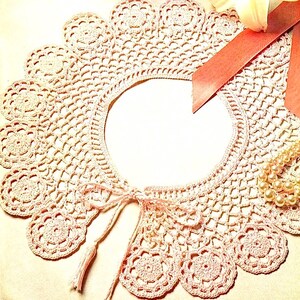 May include: A delicate crocheted collar with a scalloped edge and a bow tie. The collar is made of a light pink yarn and has a delicate, feminine look.