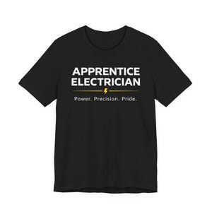 Op de afbeelding: Zwart T-shirt met de witte tekst "APPRENTICE ELECTRICIAN". Onder de tekst staat een gele bliksem en de woorden "Power. Precision. Pride." in het wit. Het T-shirt heeft een ronde hals en korte mouwen.