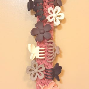 May include: A vertical macrame hair clip organizer in a dusty rose color, featuring a circular top ring and a fringed bottom. The woven fabric is adorned with multiple matte hair clips in various shapes, including flowers and butterflies in shades of pink, cream, brown, and charcoal gray.