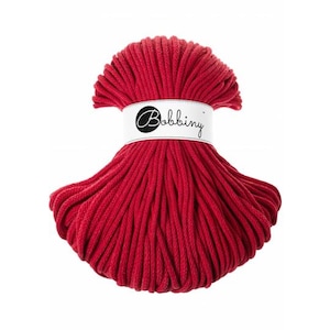 May include: A skein of deep red cotton cord for crafting, featuring a white paper band wrapped around the center. The band displays the brand name Bobbiny in a black cursive script font, with the text clearly visible against the red cord.