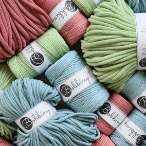 May include: Close-up of various spools of cotton rope in shades of green, blue, and pink. Each spool is labeled with the brand name "Bobbiny".
