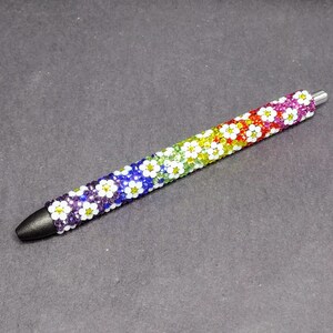 May include: A rainbow-colored pen decorated with white daisy-shaped beads. The pen has a black tip and a silver clip.