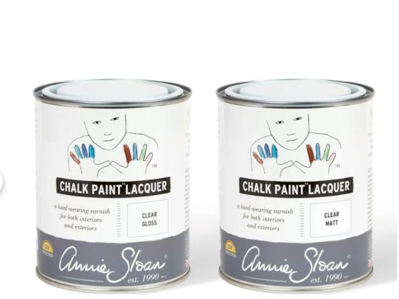 Może przedstawiać: Dwa metalowe puszki z farbą z białymi etykietami. Etykiety zawierają napis "CHALK PAINT LACQUER" i przedstawiają rysunek osoby z kolorowymi odciskami dłoni. Jedna puszka ma napis "CLEAR GLOSS", a druga "CLEAR MATT".