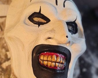 May include: A white clown mask with black accents around the eyes and mouth. The mask features a wide, menacing grin with yellow teeth and red gums. The mask is designed with a sinister expression.
