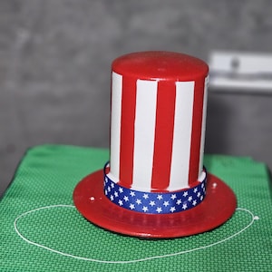 Puede incluir: Sombrero patriótico del Tío Sam con ala roja y corona cilíndrica. La corona presenta rayas rojas y blancas alternas. Una cinta azul con estrellas blancas rodea la base de la corona. El sombrero está sobre una superficie verde.