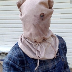 Puede incluir: Una máscara de arpillera beige con un solo agujero para los ojos y un cierre de cordón. La máscara se usa sobre una camisa a cuadros azul y un overol de mezclilla, creando una apariencia rústica e inquietante.