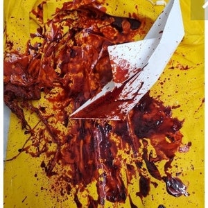 May include: A yellow surface is splattered with red liquid and a white paper boat. The red liquid appears to be blood, with droplets and larger pools. The paper boat is partially covered in the red liquid.