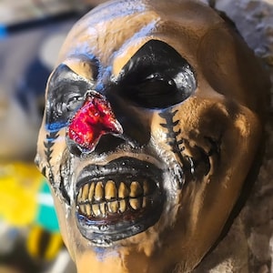 May include: A detailed, handcrafted mask with a skull-like appearance. The mask features black eye sockets, a red, textured nose, and a wide, menacing grin with yellowed teeth. The mask is painted in shades of tan and brown, with black accents.