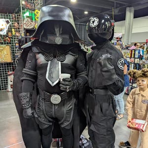 May include: Two people in black Star Wars costumes stand side-by-side. One wears a large helmet and cape, the other a helmet with a white emblem. Both are in black outfits, boots, and gloves. A child in a brown jumpsuit is in the background.