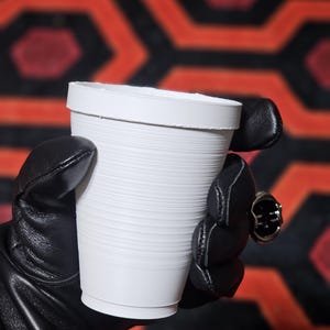 Puede incluir: Una taza de papel blanca con textura acanalada es sostenida por una persona con guantes de cuero negro. Un anillo plateado con una piedra verde oscuro es visible en la mano. El fondo presenta un patrón repetitivo de formas geométricas rojas y negras.