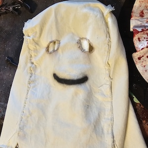May include: A cream-colored fabric mask with eye holes and a painted black smile. The edges of the mask are frayed, and the eye holes are reinforced with a tan material. The mask appears to be handmade.