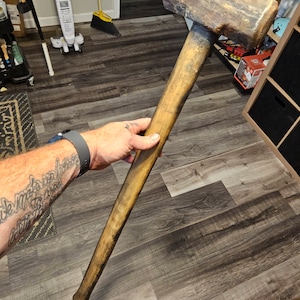 May include: A large, weathered wooden mallet with a rectangular head and a long, cylindrical handle. The head shows signs of wear and use, with a dark, aged appearance. The handle is a lighter shade of wood, with a worn finish.