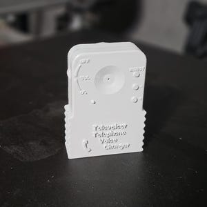 May include: A light gray 3D-printed device labeled "Televoicer Telephone Voice Changer." The device has a dial, buttons, and a speaker-like design. The text "OFF," "VOL," and "READY" are visible.