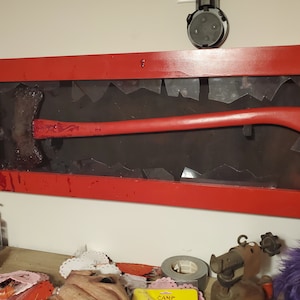 May include: A red-handled axe displayed in a red framed shadow box with a broken glass effect. The axe head is covered in a red substance. The background is black.
