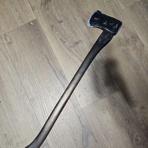 May include: Axe with a dark wooden handle and a black metal head. The axe head has a silver edge. The handle is long and curved at the end. The axe is lying on a wooden surface.