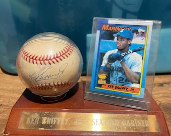May include: A baseball signed by Ken Griffey Jr. is encased in plastic, displayed next to a baseball card of the player. The card is in a protective case. The display sits on a wooden base with a brass plate that reads "Ken Griffey Jr. Seattle Mariners."