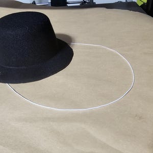 May include: A black top hat with a rounded crown and a wide brim. The hat is positioned on a light brown surface, with a white circular outline drawn around it. The hat appears to be made of a textured fabric.