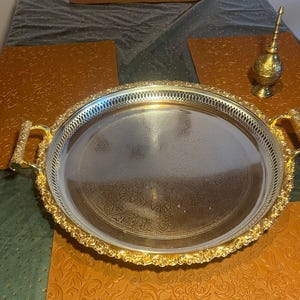 Può includere: Un vassoio argentato con manici dorati ornati e un bordo decorativo dorato. Il vassoio presenta un disegno inciso all'interno. Un piccolo contenitore decorativo dorato con un lungo beccuccio sottile è nelle vicinanze.
