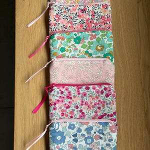May include: A stack of six small, rectangular floral print pouches with pink zippers and matching ribbon pulls. The pouches feature various floral patterns in shades of pink, blue, yellow, and green, set against white and pastel backgrounds.