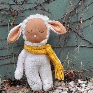 Puede incluir: Un juguete de peluche de cordero blanco con una cara marrón y los ojos cerrados. El cordero lleva una bufanda amarilla y está sentado en una cama de hojas marrones y piedras.