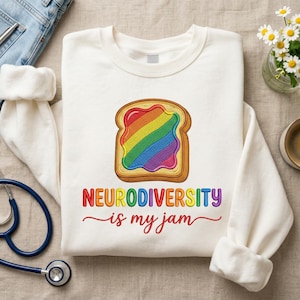 Pode incluir: Camisola creme com um gráfico colorido de torrada com geleia arco-íris e o texto "NEURODIVERSITY is my jam". Um estetoscópio, jeans e uma chávena de café também estão na imagem.