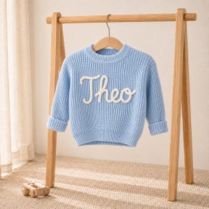 Pode incluir: Um suéter de malha de manga comprida em azul claro com gola redonda, punhos e bainha canelados. A parte frontal do suéter exibe o nome Theo em letras cursivas brancas grandes e em relevo. A peça está pendurada em um cabide de madeira.