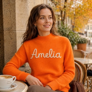 May include: A bright orange long-sleeve sweater featuring a mock neck collar and the name Amelia embroidered in white cursive script across the chest. The knit garment has ribbed cuffs and a ribbed hem, providing a textured appearance.