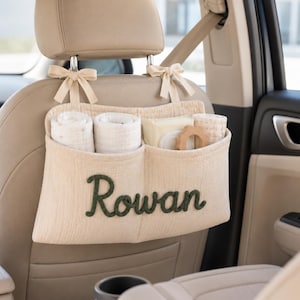 Peut inclure: Un organisateur de siège auto en tissu beige doté de deux compartiments principaux et de deux rubans pour la fixation à l'appui-tête. L'avant est brodé du nom Rowan en écriture cursive vert foncé. Il contient des tissus roulés et un anneau.
