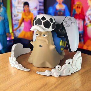 May include: A tan and blue character-shaped gaming controller stand with a white and black hat. A white gaming controller rests on the stand. White decorative accents are on the table. The background is blurred with colorful images.