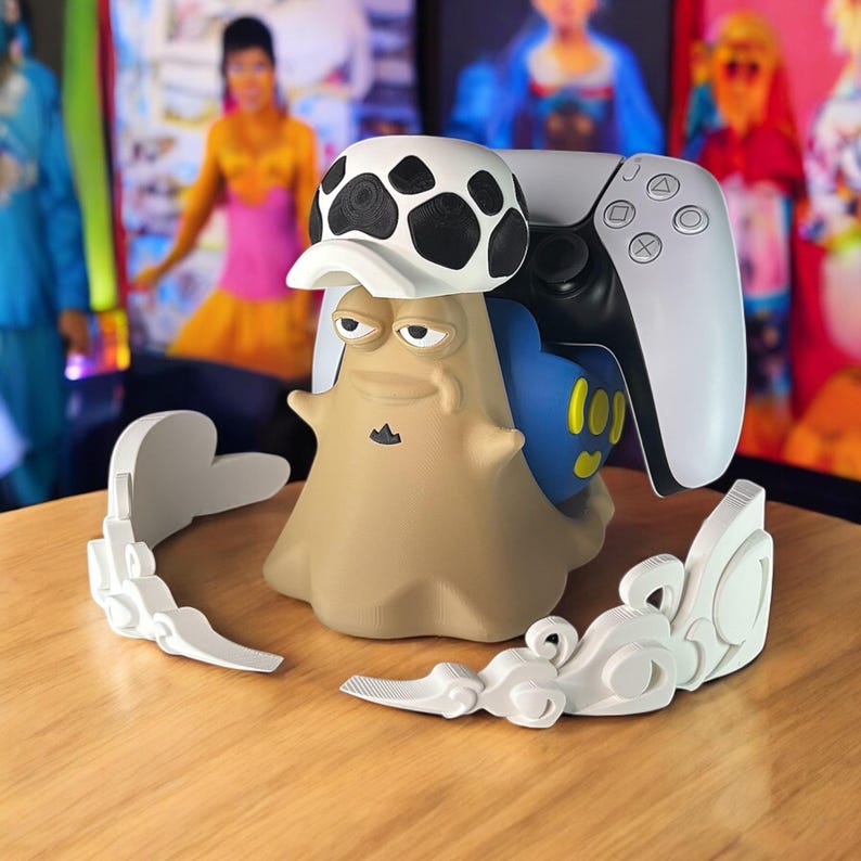 May include: A tan and blue character-shaped gaming controller stand with a white and black hat. A white gaming controller rests on the stand. White decorative accents are on the table. The background is blurred with colorful images.
