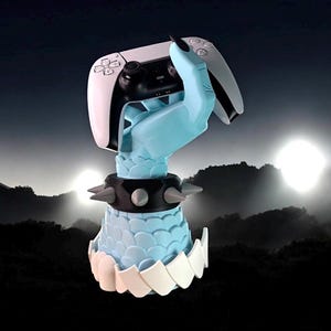 K&ouml;nnte beinhalten: Ein hellblauer Gaming-Controller-St&auml;nder in Form einer Hand. Der St&auml;nder hat ein schwarzes Armband mit Nieten und eine schuppenartige Basis. Ein wei&szlig;er und schwarzer Gaming-Controller wird in der Hand gehalten. Der St&auml;nder ist f&uuml;r einen Gaming-Controller konzipiert.