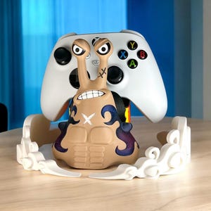May include: A tan and purple video game controller stand shaped like a cartoon character, with a white Xbox controller resting on top. The stand has a smiling face and is surrounded by white cloud-like accents.