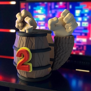 Peut inclure: Un récipient en forme de tonneau marron avec un grand chiffre 2 rouge et jaune sur le côté. Deux poings blancs émergent du dessus, avec une petite tasse blanche entre eux. Le récipient a un aspect boisé texturé.