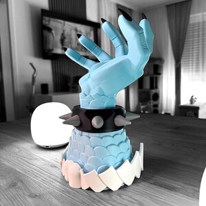K&ouml;nnte beinhalten: Hellblaue, stilisierte Handskulptur mit schwarzen Fingern&auml;geln. Die Hand tr&auml;gt einen schwarzen Kragen mit silbernen Stacheln und Nieten. Der Sockel ist hellblau mit wei&szlig;-rosa Zierleiste. Die Skulptur steht auf einer Holzoberfl&auml;che.