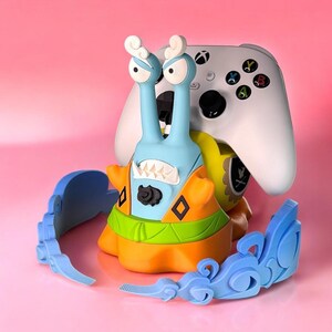 May include: A blue and orange snail-shaped video game controller stand with a white Xbox controller on top. The stand has a green and yellow shell and a black face with a toothy grin. The stand is sitting on a pink surface with a blue decorative piece.