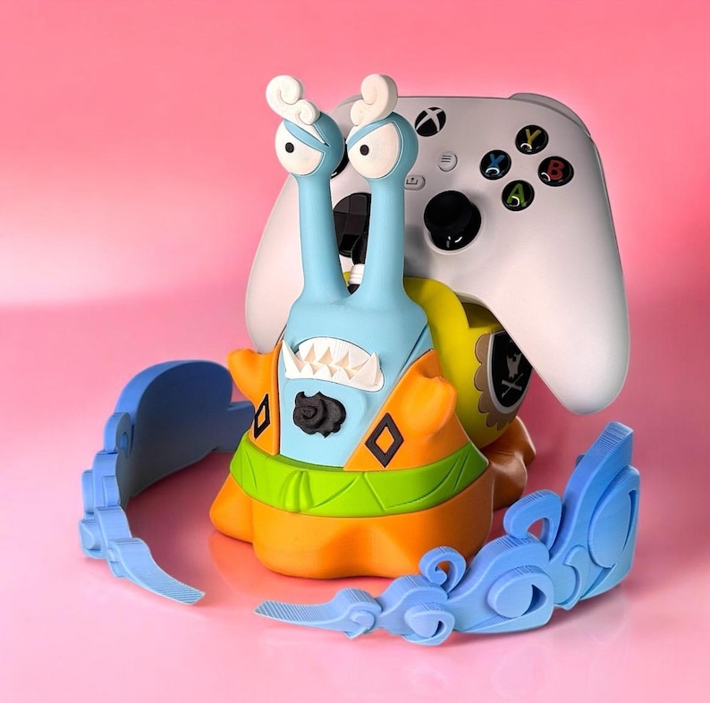 May include: A blue and orange snail-shaped video game controller stand with a white Xbox controller on top. The stand has a green and yellow shell and a black face with a toothy grin. The stand is sitting on a pink surface with a blue decorative piece.