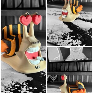 May include: A whimsical 3D-printed snail figurine with a beige body, a striped shell, and heart-shaped eyes. The snail has an open mouth with visible teeth and a small object. The figurine is set against a black and white background. The text "3D FACTORY BOX" is visible.