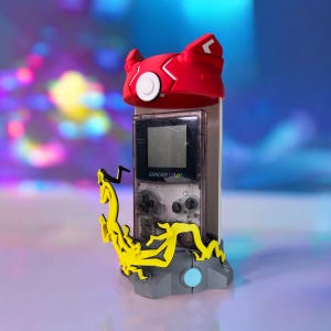 K&ouml;nnte beinhalten: Ein grauer 3D-gedruckter St&auml;nder mit einem gelben Drachen-Design h&auml;lt einen Game Boy Color mit einer roten und wei&szlig;en Pokemon-Charakterkappe oben drauf. Der Bildschirm des Game Boy Color ist schwarz und die Kn&ouml;pfe sind grau.