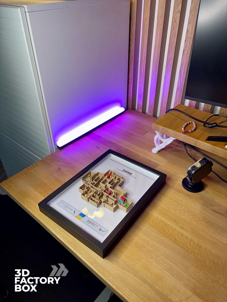 May include: A framed 3D model of a building layout, labeled "CACHE", with miniature figures and colored markers. The frame is black, and the model is set on a wooden desk. The logo "3D FACTORY BOX" is in the lower left corner.