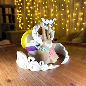 Pode incluir: Uma figura de caracol castanho com um rosto sorridente, olhos roxos e uma estrela rosa de um lado. O caracol tem uma concha amarela e roxa e detalhes brancos. A figura está sobre uma superfície de madeira.