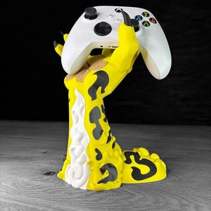 K&ouml;nnte beinhalten: Eine gelbe und schwarze Monsterhand h&auml;lt einen wei&szlig;en Videospiel-Controller. Die Hand h&auml;lt den Controller in einem krallenartigen Griff. Der Controller ist ein Standard-Xbox-Controller.