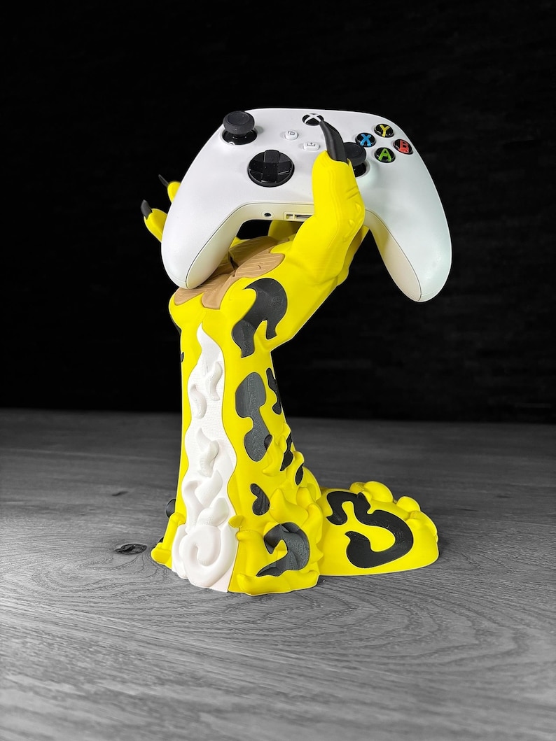 K&ouml;nnte beinhalten: Eine gelbe und schwarze Monsterhand h&auml;lt einen wei&szlig;en Videospiel-Controller. Die Hand h&auml;lt den Controller in einem krallenartigen Griff. Der Controller ist ein Standard-Xbox-Controller.