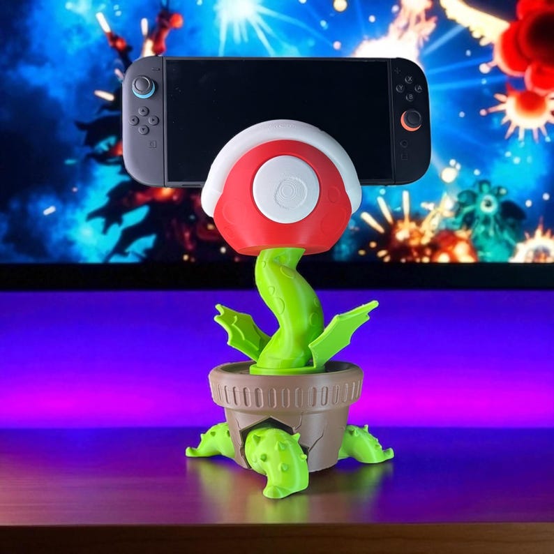May include: A novelty plant-shaped phone or game console holder. The holder features a red and white flower head, green stem and leaves, and a brown pot base. A gray Nintendo Switch console is placed on top of the flower.