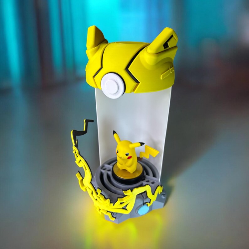 May include: A yellow Pikachu Pokemon figure is inside a clear plastic container with a yellow lid. The container has a gray base with a lightning bolt design.