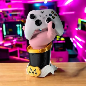 May include: A white Xbox controller held by a pink hand-shaped stand. The stand is black with a gold trim and a gold emblem. The background is a blurred gaming setup with neon lights.