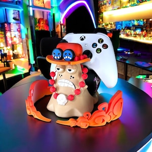 Könnte beinhalten: Ein 3D-gedruckter Controller-Ständer mit einer Comicfigur mit einem rot-weißen Hut, einem braunen Körper und roten Perlen. Der Ständer ist für zwei weiße Gamecontroller ausgelegt.