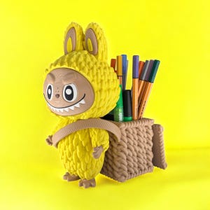 May include: A whimsical yellow pen holder shaped like a creature with a bunny-like hood and a basket on its back. The pen holder is filled with colorful pens and markers, set against a bright yellow background.